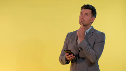 Young businessman in suit looking on smartphone and thinking about something on yellow background. He probably making some plans.