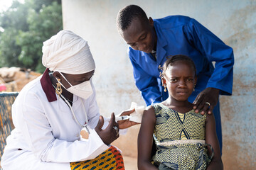 Frightened little black African girl being vaccinated by a female nurse with face mask, with a man in a blue coat bent over her patting her shoulder reassuringly