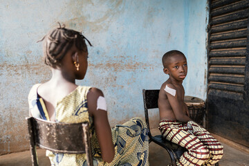 Two black siblings sitting outside a rural dispensary with big plasters on their upper arms after a vaccination in an African village