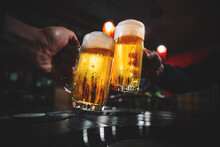 Two Glass Of Beer In Hand. Beer Glasses Clinking In Bar Or Pub