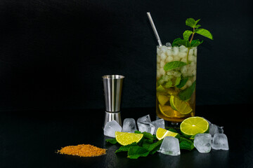 Mojito cocktail with fresh peppermint, lime, ice cubes and meter bar, brown sugar with black slate background.