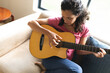 © Wavebreak Media - Mixed race woman sitting on sofa and playing guitar