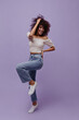 © Look! - Excited brunette curly woman in jeans and cropped white top moves, smiles, touches hair and rises leg on purple background.
