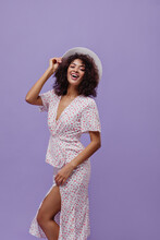 Cheerful Curly Woman In White Polka Dot Dress Smiles On Purple Background. Happy Dark-skinned Lady Holds Hat On Isolated.