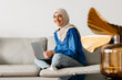 Middle eastern woman in headscarf using laptop while sitting on sofa