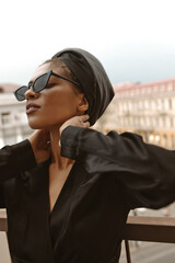 Wall Mural - Beautiful lady in cool sunglasses looking away on balcony. Young woman in long sleeve black clothes and unusual hat posing on terrace..