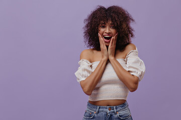 Wall Mural - Surprised curly woman looks into camera on isolated. Happy brunette dark-skinned lady in white top poses on purple background.