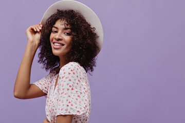 Wall Mural - Charming brunette woman in white hat and floral dress smiles sincerely, looks into camera and poses on purple background.