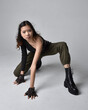 Full length portrait of pretty brunette, asian girl wearing black top and khaki utilitarian army pants and leather boots. Sitting pose , isolated agent a light grey studio background.