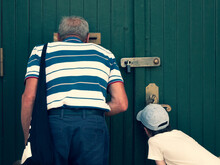 Senior And Child Are Looking Through The Mail Slot And Keyhole, View From The Back. Grandfather And Grandson See Something Interesting. Concept Of Two Generations, Peeping And Peeking