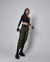 Full length portrait of pretty brunette, asian girl wearing black top and khaki utilitarian army pants and leather boots. Standing pose with gestural hands, isolated against a light grey studio backgr