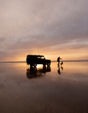 Travel Lifestyle View Of Land Rover Defender Driving On Beach During Golden Sunset With Person And Dog, Black Rock Sands, Near Snowdonia, North Wales.