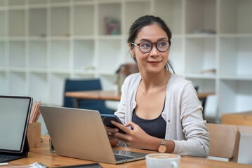 Wall Mural - Portrait of young Asian businesswoman wearing glasses holding a smartphone a laptop placed at the office.