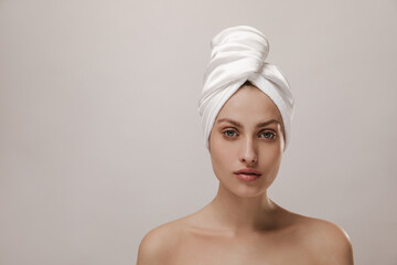 Close-up portrait of charming young lady with pure skin and white towel on head, looking into camera isolated over light background. Skin care and health concept