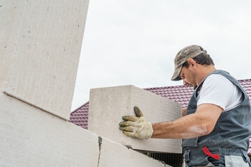 young courageous male construction worker in overalls is laying aerated concrete blocks on a construction site. High-quality laying of blocks on glue, building a house.. Copy space