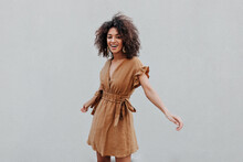 Excited Dark-skinned Lady In Beige Dress Smiles And Turns On Grey Background. Curly Brunette Woman In Brown Outfit Poses Near Wall.