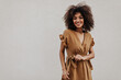 © Look! - Young African woman in brown dress looks into camera. Dark-skinned curly lady in stylish outfit smiles on grey background.