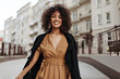 © Look! - Brunette dark-skinned woman in brown dress and black coat walks outdoors. Curly lady smiles sincerely and moves outside.