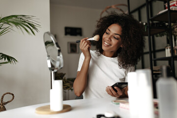 Wall Mural - Joyful young brunette woman in white t-shirt smiles, powders face, holds palette abc makeup brush, looks into mirror.