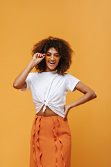 Wall Mural - Positive girl in sunglasses smiles on isolated backdrop. Curly lady in white t-shirt and and orange skirt posing on yellow background..