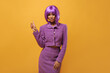 Sad woman in purple short wig shows to place for text on yellow background. Stylish lady in lilac warm jacket and skirt posing on isolated backdrop..