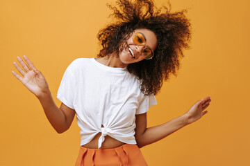 Wall Mural - Wonderful woman in white t-shirt and bright sunglasses smiled on isolated backdrop. Dark-skinned girl with curly hair has fun on yellow background..