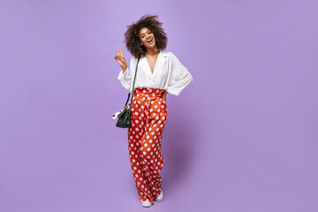 Wall Mural - African lady in wide polka dot pants shows peace sign on lilac background. Brunette woman in blouse and handbag on isolated backdrop..