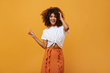 Optimistic lady in white t-shirt and orange skirt smiling on yellow background. Curly girl looks into camera on isolated backdrop..