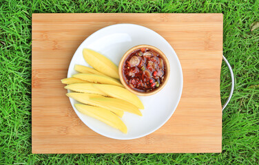 Wall Mural - Sliced Mango sour with sweet fish sauce in white plate over bamboo wooden board against green grass background. Top view