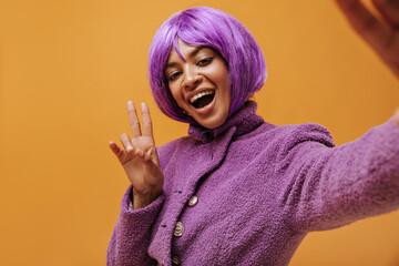 Wall Mural - African woman in wig and lilac warm jacket making selfie on isolated backdrop. Happy girl with short hair shows peace sign on orange background..