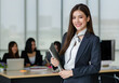 © Bangkok Click Studio - Portrait of young attractive Asian female office worker in formal business suits  smiling at camera in office with blurry colleagues sitting in office as background