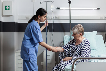 Medical nurse helping senior woman patient getting up from bed in hospital room, with iv drip bag attached while she gets intravenous treatment and oxymeter measuring blood oxygen saturation.