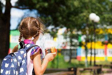 Girl with a backpack take off the medical mask and eating pie near the school. A quick snack with a bun, unhealthy food, lunch from home. Back to school. Education, primary school classes, September 1