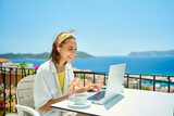 Fototapeta  - beautiful caucasian woman in wireless earphones talk speak on video call on computer while sitting at balcony with seaview on resort. female have online conversation, work chat conference or