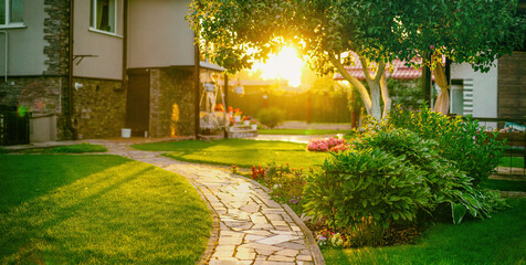 Fotomurales - Beautiful manicured lawn and flowerbed with deciduous shrubs on private plot and track to house against backlit bright warm sunset evening light on background. Soft focusing in foreground.