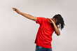 Portrait of young adult man with dreadlocks wearing red casual style T-shirt, standing in dab dance pose, internet meme, celebrating success. Indoor studio shot isolated on gray background.