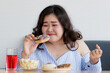 © Bangkok Click Studio - Happiness closeup shot of Asian young happy beautiful hungry overweight fat chubby female smile at camera hold chocolate donut in hand and hold fist up satisfy with dessert cake popcorn on table