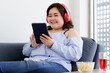 © Bangkok Click Studio - Asian young happy pretty friendly fat plus size woman sit smiling relaxing on gray fabric couch listening to music from big red headphones use playlist from black tablet on hand in living room