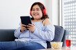 © Bangkok Click Studio - Asian young happy pretty friendly fat plus size woman sit smiling relaxing on gray fabric couch listening to music from big red headphones use playlist from black tablet on hand in living room