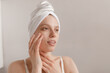© Look! - Gorgeous lady applying cream on pure skin. Girl with white towel wrapped hair, making procedures and looking aside isolated over light background. Cosmetology and skincare concept