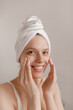 © Look! - Pretty pale girl making procedures. Lady with white towel wrapped hair smiling and applying cream on pure face and looking into camera against light background