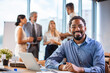 © Dragana Gordic - Businessman Using Laptop At Desk In Busy Office.  Black businessman with colleagues sitting in a modern board room. Proud smiling business man sitting during a meeting and looking at camera
