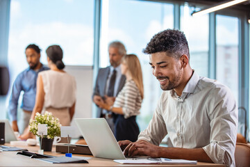 Wall Mural - Businessman Using Laptop At Desk In Busy Office.  Black businessman with colleagues sitting in a modern board room. Proud smiling business man sitting during a meeting and taking notes