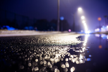 Wall Mural - Foggy night in the big city, the street lighting lanterns and headlights of the approaching car. Close up view from the level of the puddle on the pavement