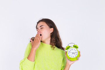 Young beautiful woman with freckles light makeup in sweater on white background with alarm clock angry tired sleepy exhausted yawning