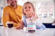 Father And Daughter Putting Pocket Money Into Savings Jar On Kitchen Counter