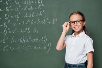cheerful schoolchild adjusting eyeglasses near chalkboard with mathematic equations
