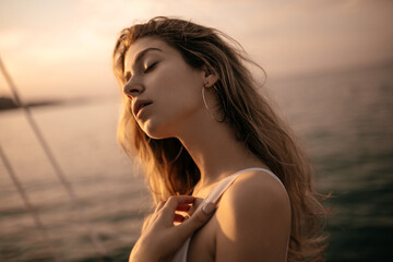 Wall Mural - Portrait of tender young lady with blonde fluffy hair, closed eyes in white clothes and round earrings against sunlit sea and sky background during beautiful pastel pink sunset