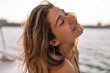 © Look! - Portrait of cheerful young lady with fluffy blonde hair, closed eyes and smile on face against background of sunlit blue sea
