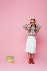 Wall Mural - shocked girl touching head and looking at camera near stack of textbooks on pink background